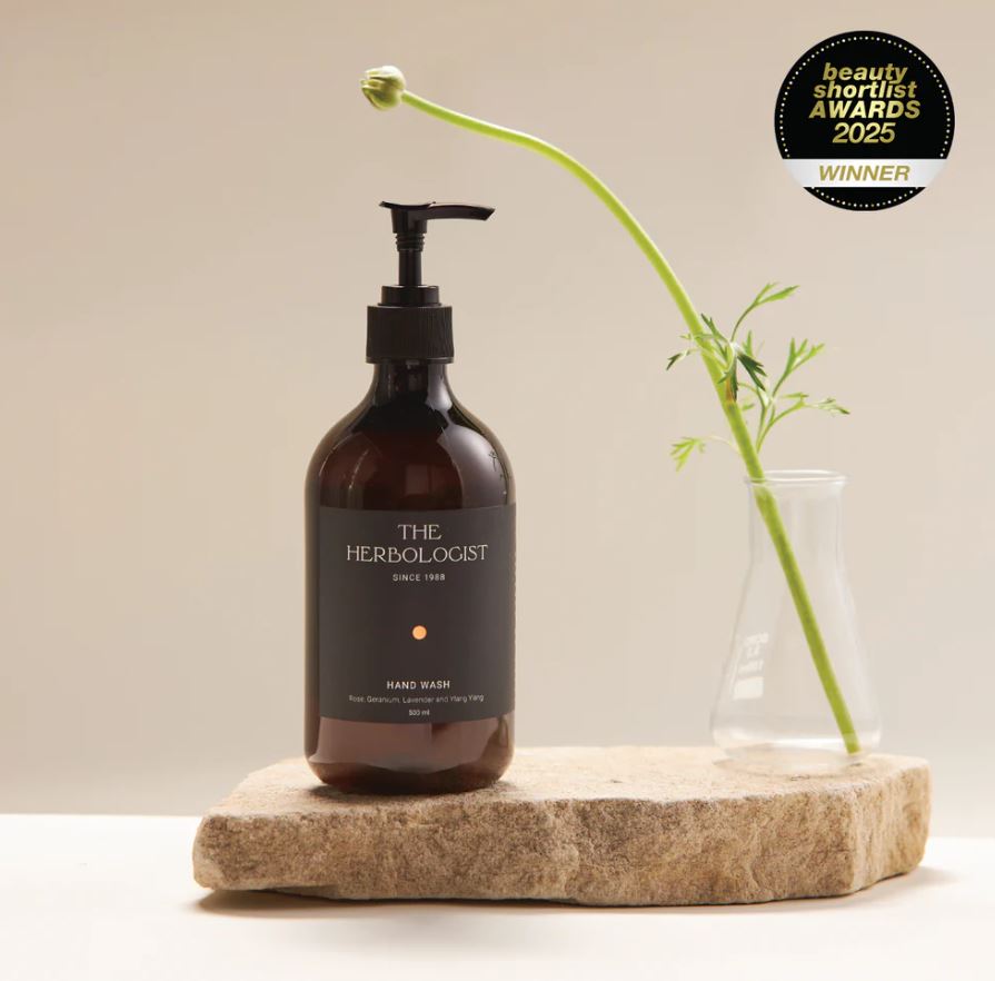 Hand wash bottle labeled 'The Herbologist' on a wooden block with a plant, against a beige background.