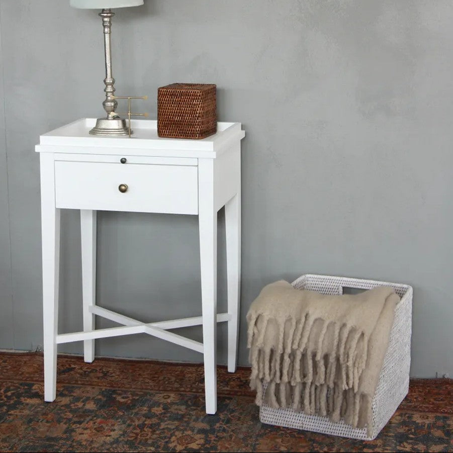 White side table with lamp and basket against a gray wall, next to a wicker basket with a blanket.